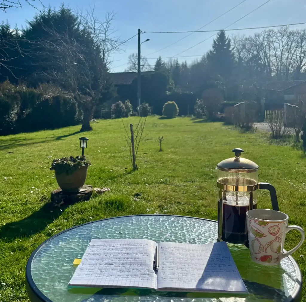 A notebook, coffee pot and mug on a garden table with the garden in the background.
